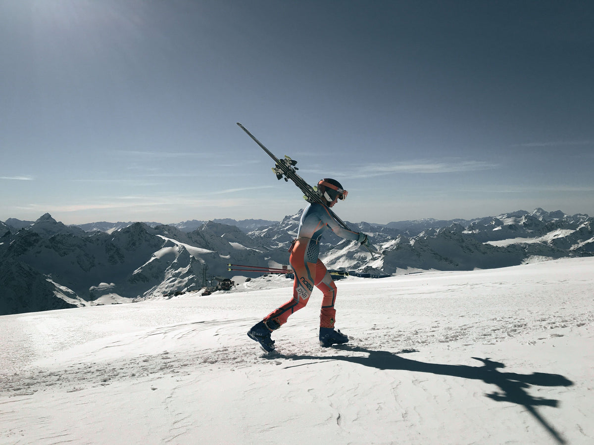 Skier carrying skis across a snowy summit with mountains behind