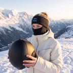 Illustrative image (AI-generated): skier on a snowy ridge wearing the Tunes Band in Black over a balaclava, helmet in hand